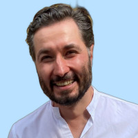 A man with a beard and wavy hair with flecks of gray. He’s wearing a very casual white shirt against a blue-sky background.